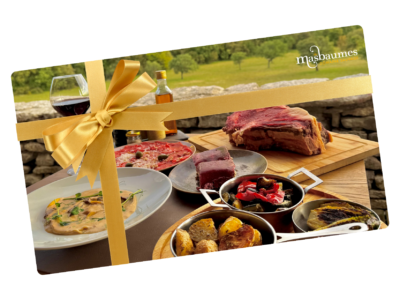 Festive gourmet spread on a wooden table with a large golden ribbon and bottle of wine outdoors, suggesting a gift hamper.