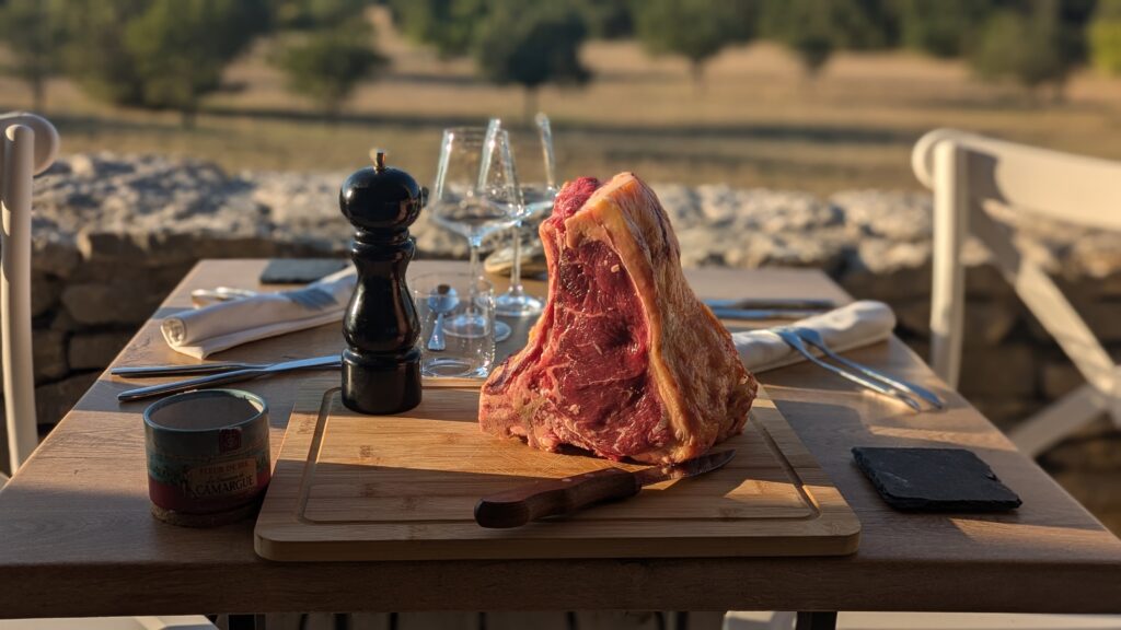 Outdoor dining setup with a large raw rib roast on a cutting board, salt shaker, knife, and wine glasses nearby.