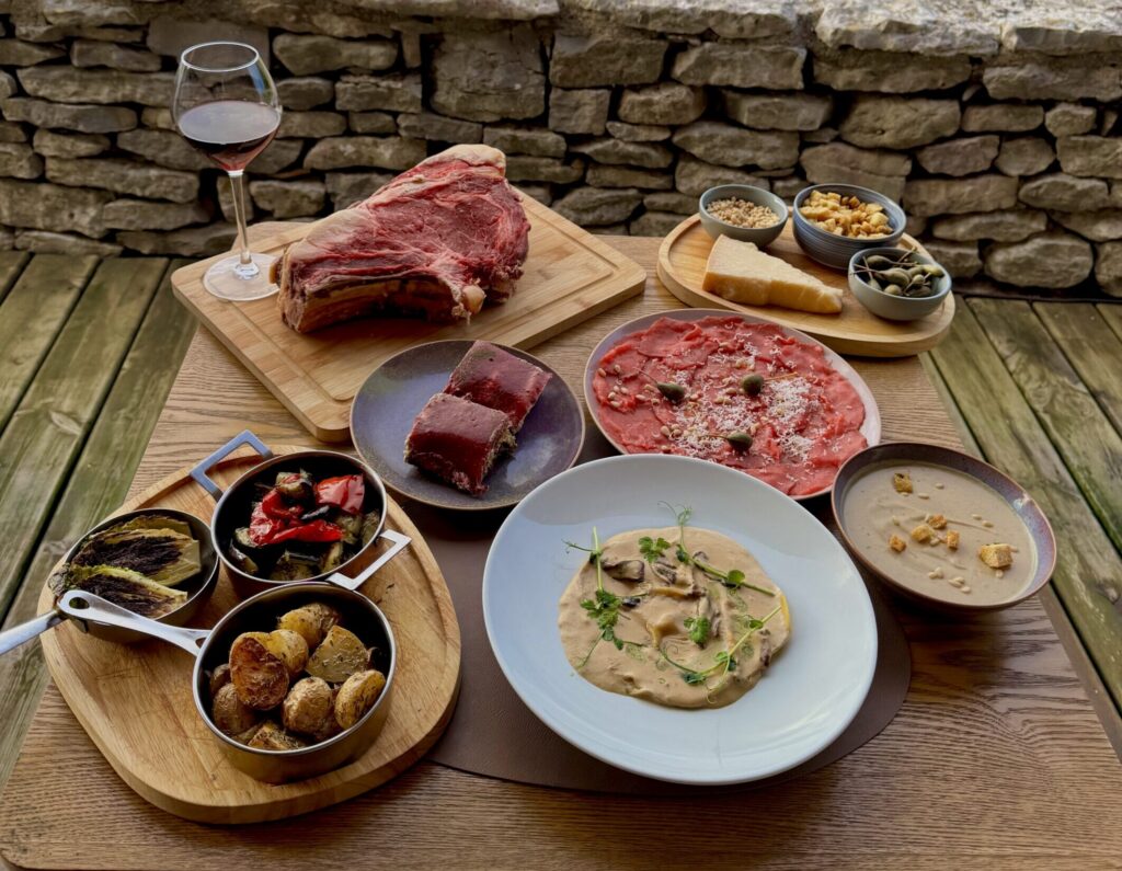 Outdoor rustic table spread with meat, dips, and bread on wooden boards, glass of red wine, against a stone wall and green field with trees in the background