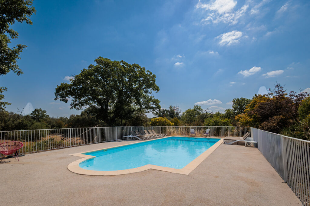 Outdoor swimming pool area with lounge chairs along a white fence and blue sky above. Trees line the background.