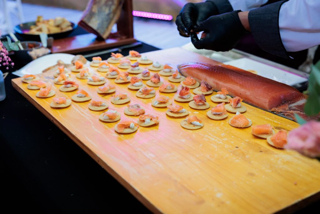 Small round crackers topped with smoked salmon and cream cheese arranged on a wooden board at a buffet.