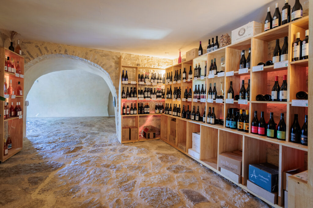 Wine cellar with wooden shelves lined with wine bottles, stone walls, and an arched doorway in the background.