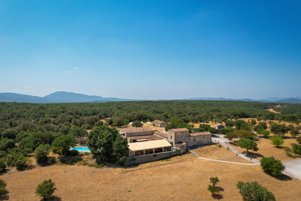 Rural village with stone houses surrounded by trees, open fields, and distant blue hills under a clear sky.]