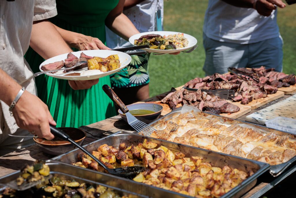 Outdoor barbecue scene with guests serving plates of sliced steak, roasted potatoes, and vegetables from long trays.