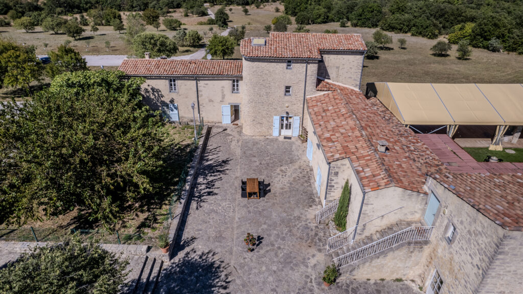Aerial view of a beige stone building complex with red-tiled roofs, blue doors, and a cobblestone courtyard surrounded by trees and fields.