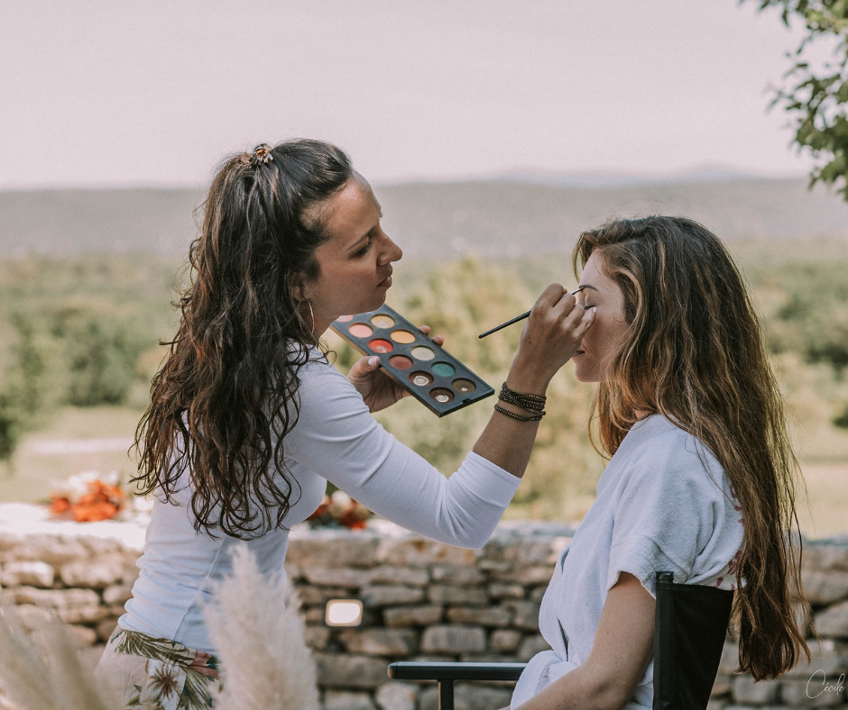 Makeup artist applies eye makeup to a seated woman outdoors, using an colorful palette on a sunny day.