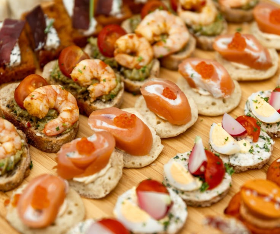 Assorted canapés on a wooden board, including shrimp-topped crostini, smoked salmon on crackers, and mini deviled eggs.